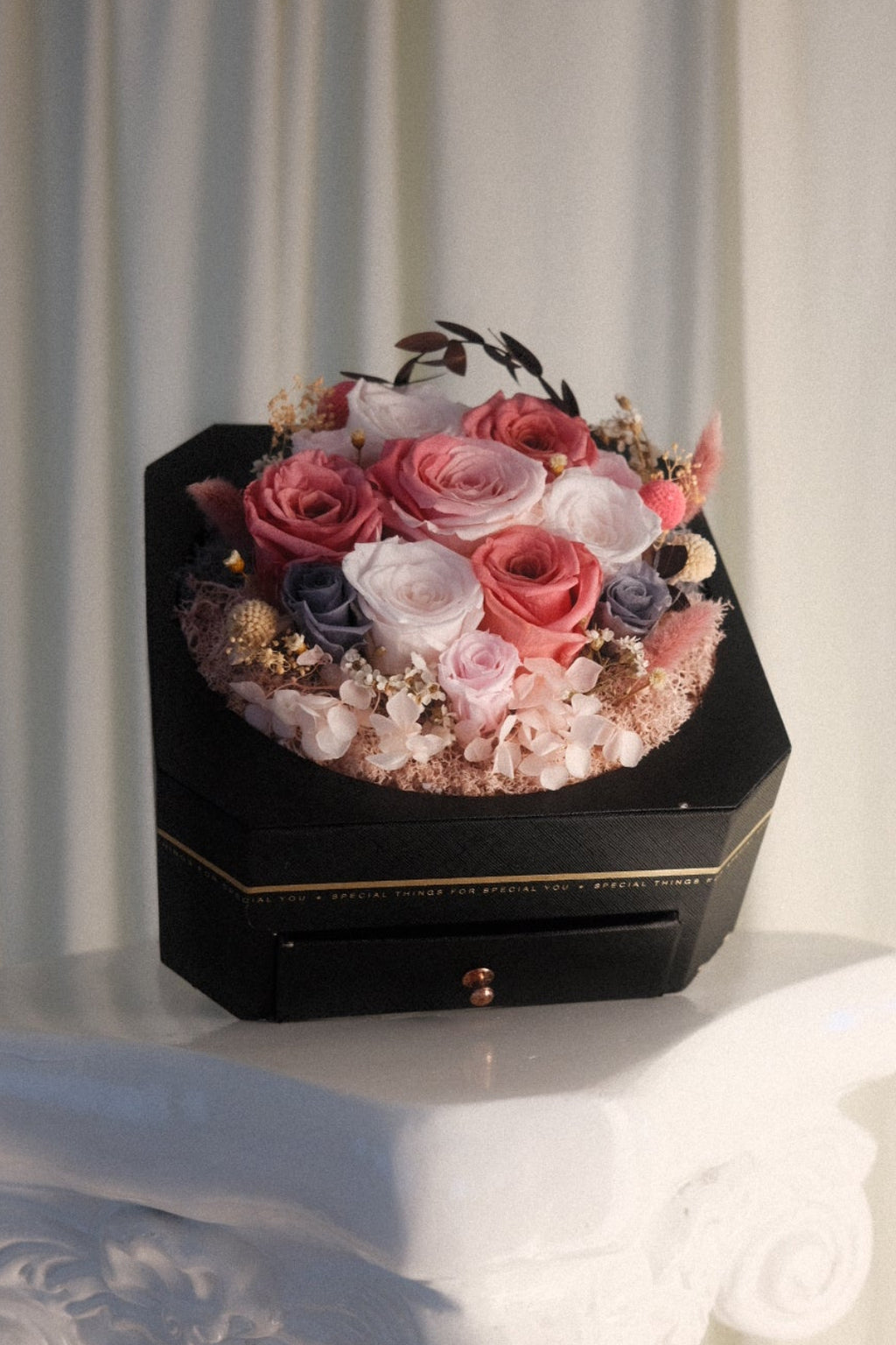 A black hexagonal flower box filled with pink, red, and white roses, placed on a white decorative pedestal in front of a soft beige curtain backdrop.