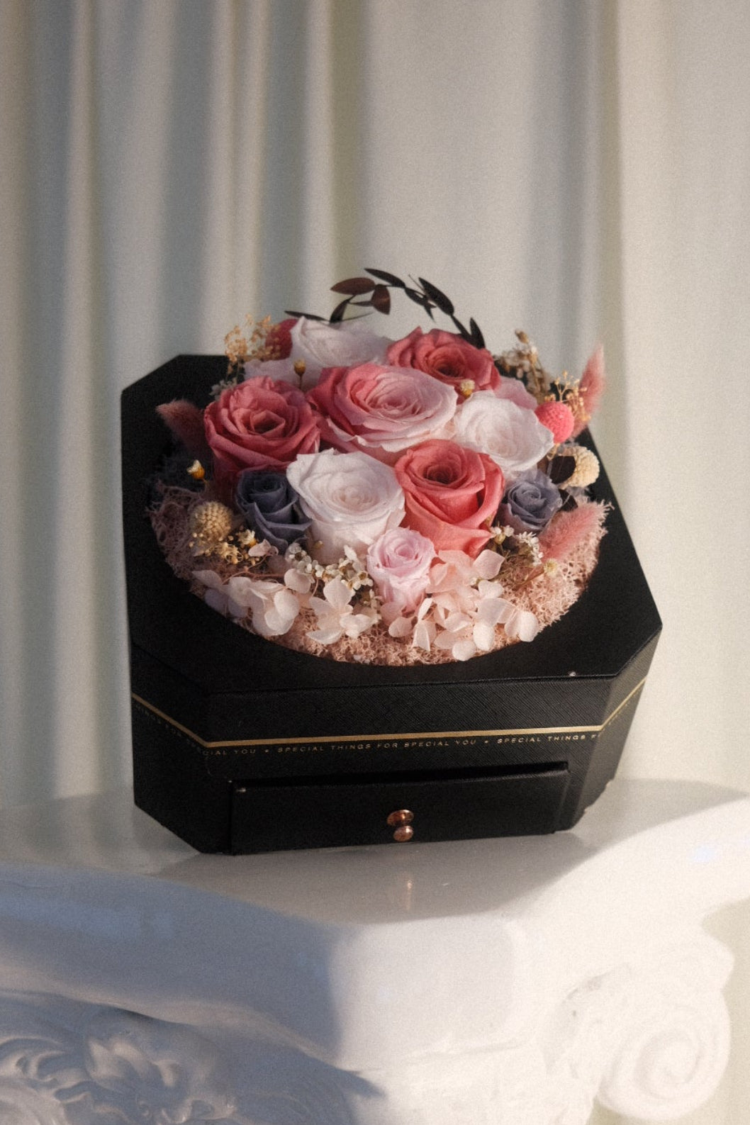 A black hexagonal flower box filled with pink, red, and white roses, placed on a white decorative pedestal in front of a soft beige curtain backdrop.