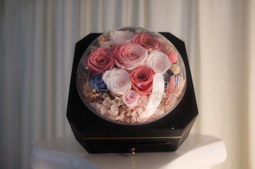 A preserved flower arrangement in a clear dome display resting on a black octagonal box, set against a soft cream curtain background.