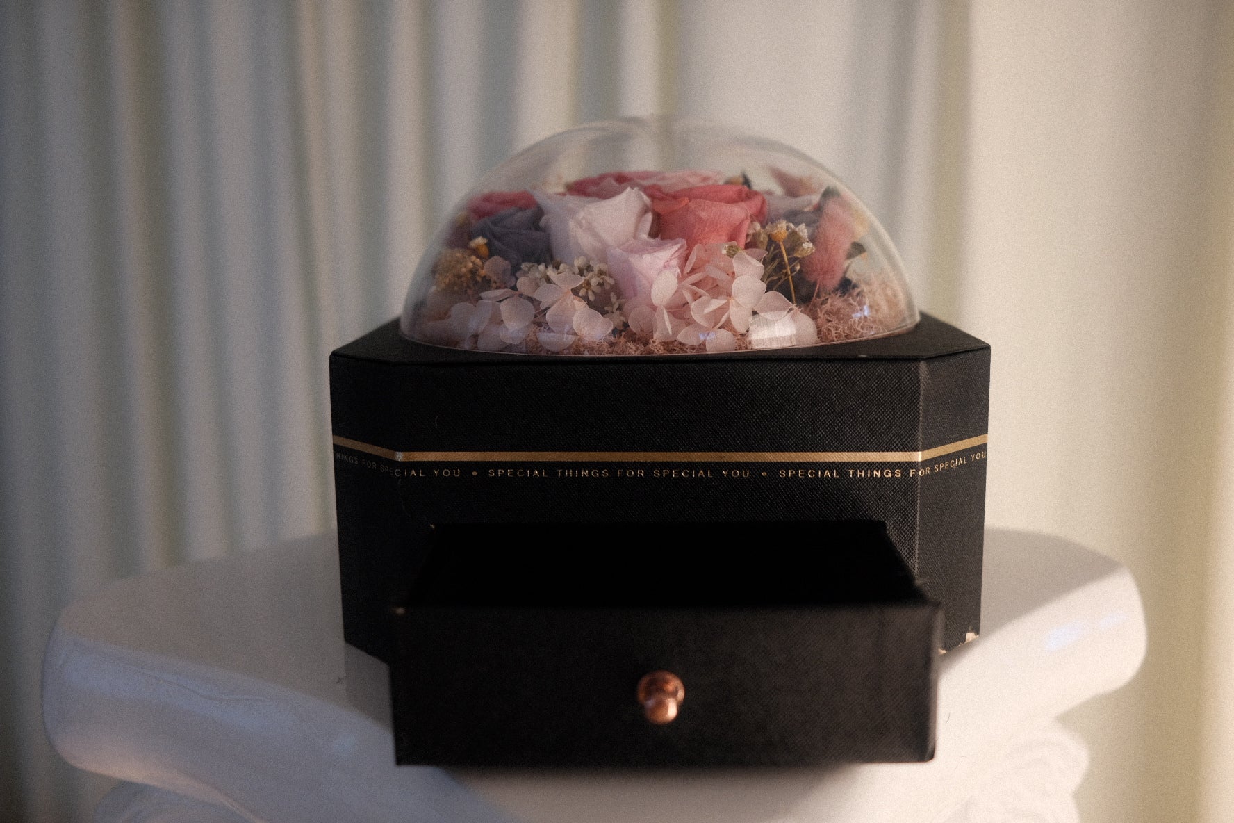A black square gift box with a single front drawer and a clear dome lid, filled with dried pink and white flowers. The box sits on a white pedestal against a soft beige curtain background.