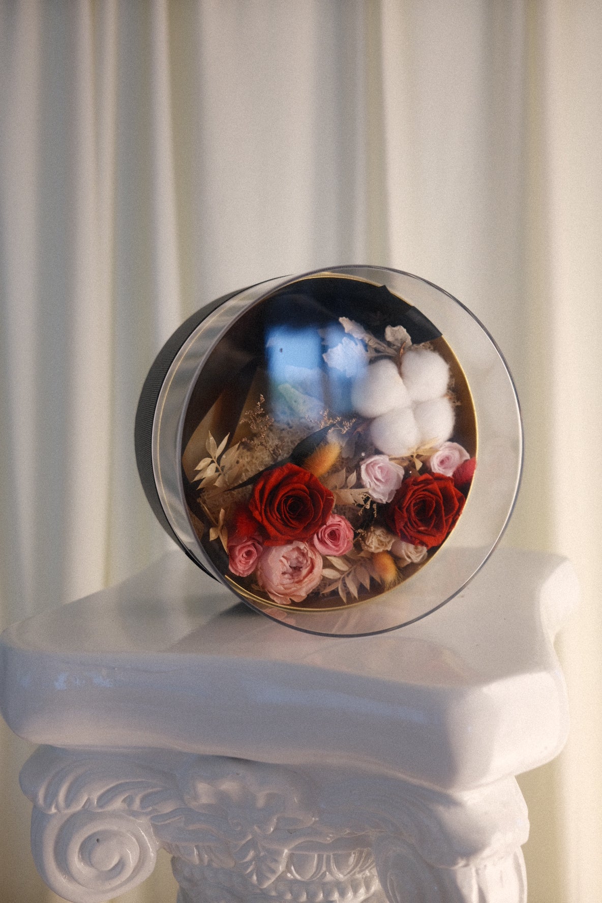 A round glass preserved gift box filled with preserved flowers, cotton bolls, and small botanical elements, displayed on a white decorative pedestal against a soft cream curtain background.