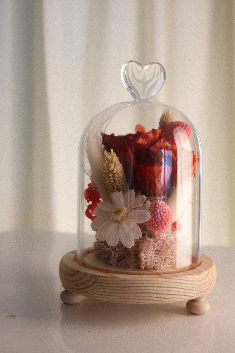 A glass cloche dome with a heart-shaped top handle, showcasing a dried flower arrangement in red, pink, and white tones, resting on a light wood base with small feet, set against a soft cream curtain background.