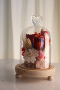 A glass cloche dome with a heart-shaped top handle, showcasing a dried flower arrangement in red, pink, and white tones, resting on a light wood base with small feet, set against a soft cream curtain background.