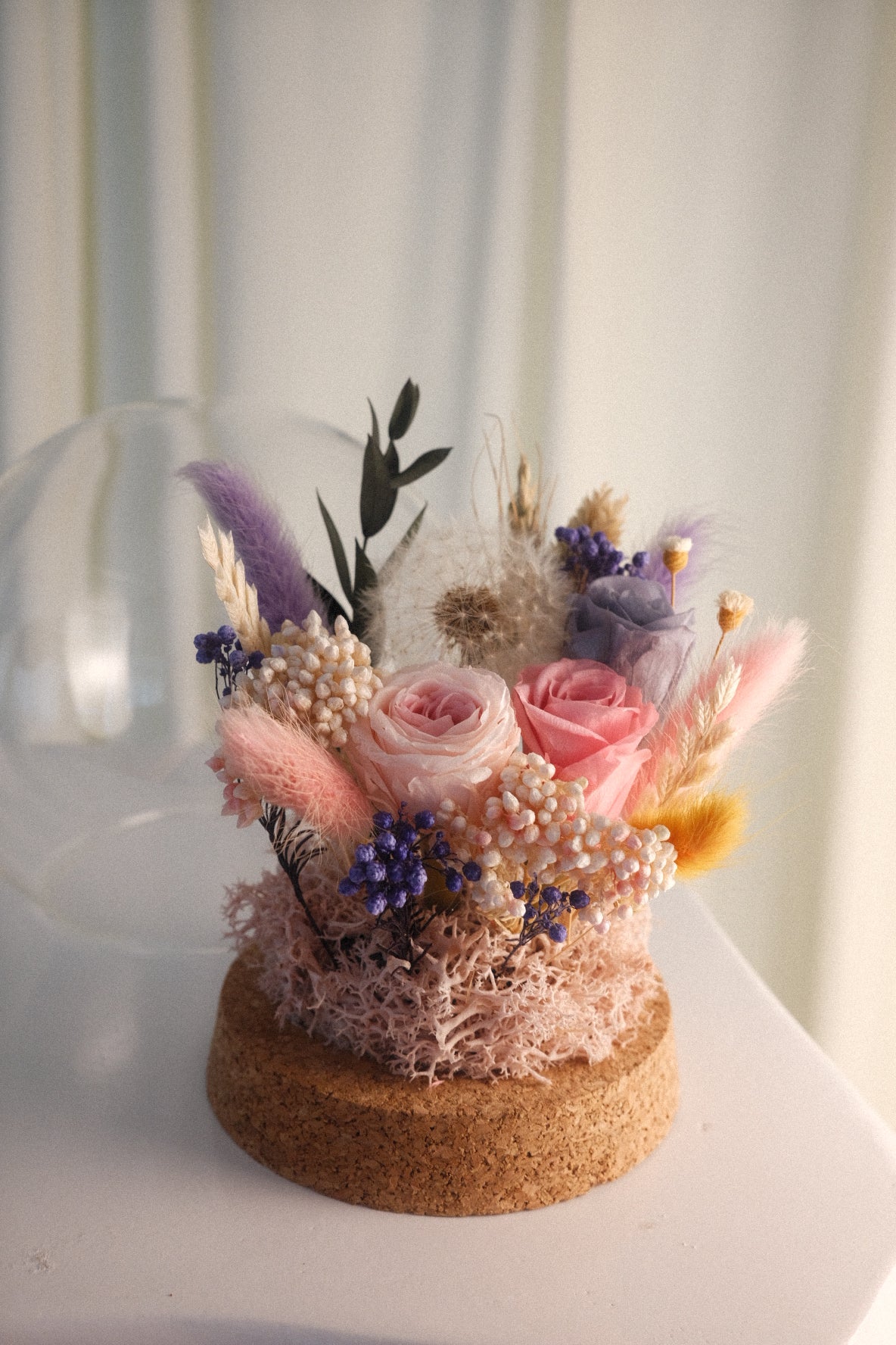 A dried flower arrangement in a round cork base, featuring pink roses, lavender pampas grass, purple baby's breath, and white pearl-like accents, displayed on a white surface with a soft neutral background.