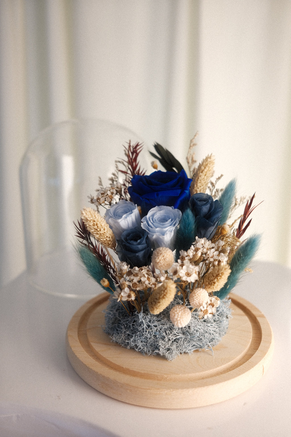 A preserved floral arrangement featuring blue and navy roses with dried grasses, small cream blossoms, teal feathers, and moss, displayed on a round wooden base with a glass dome cover in the background.
