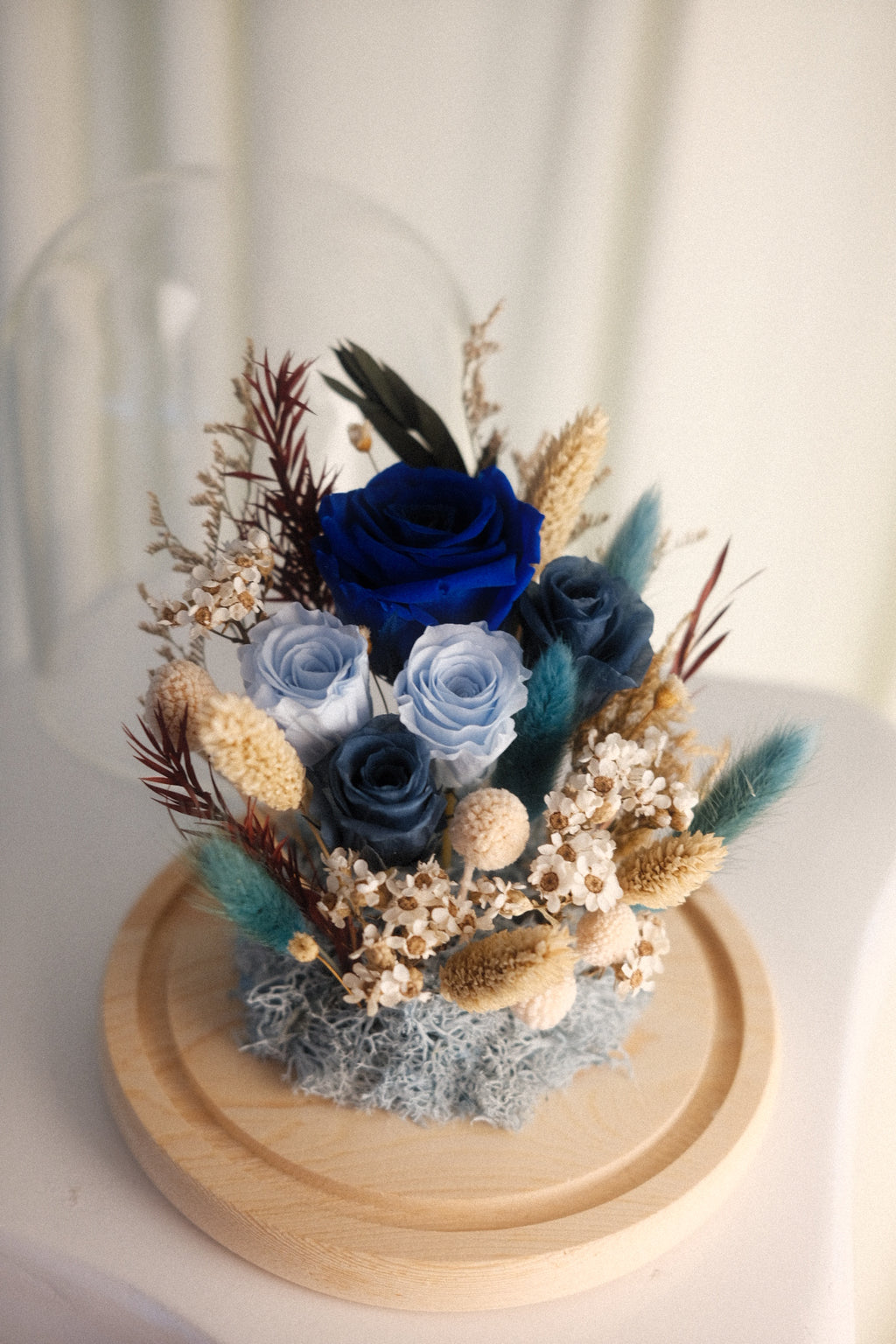 A preserved floral arrangement in a round wooden display base, featuring a prominent deep blue rose with light blue and navy roses, dried grasses, and teal accents, under a clear glass dome.
