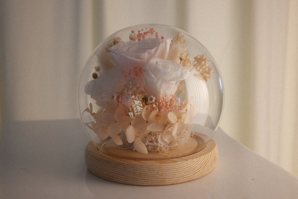 A clear glass dome snow globe containing dried white and pale pink flowers, resting on a round light wood base, set on a white surface against a soft beige curtain background.