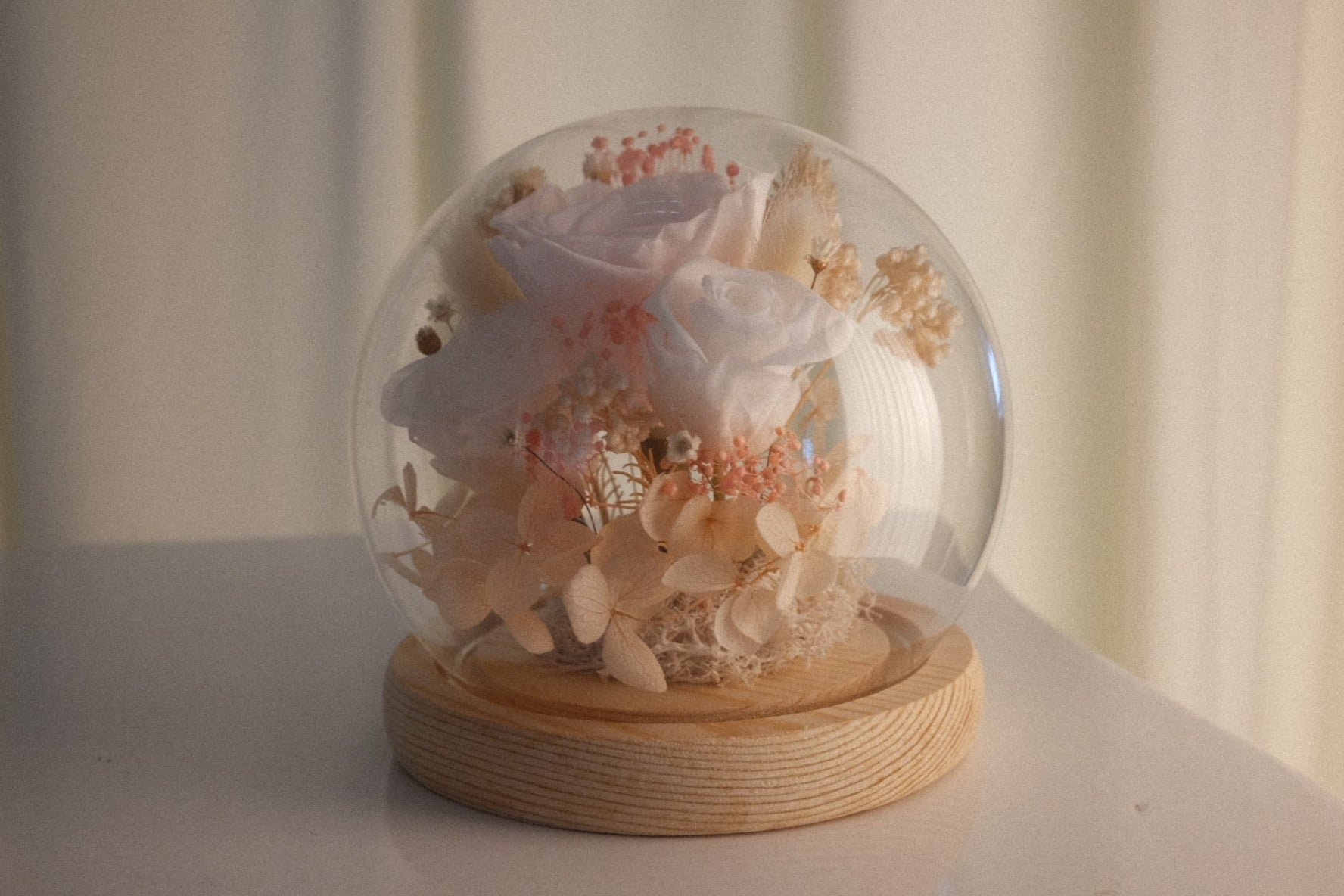 A clear glass dome snow globe containing dried white and pale pink flowers, resting on a round light wood base, set on a white surface against a soft beige curtain background.