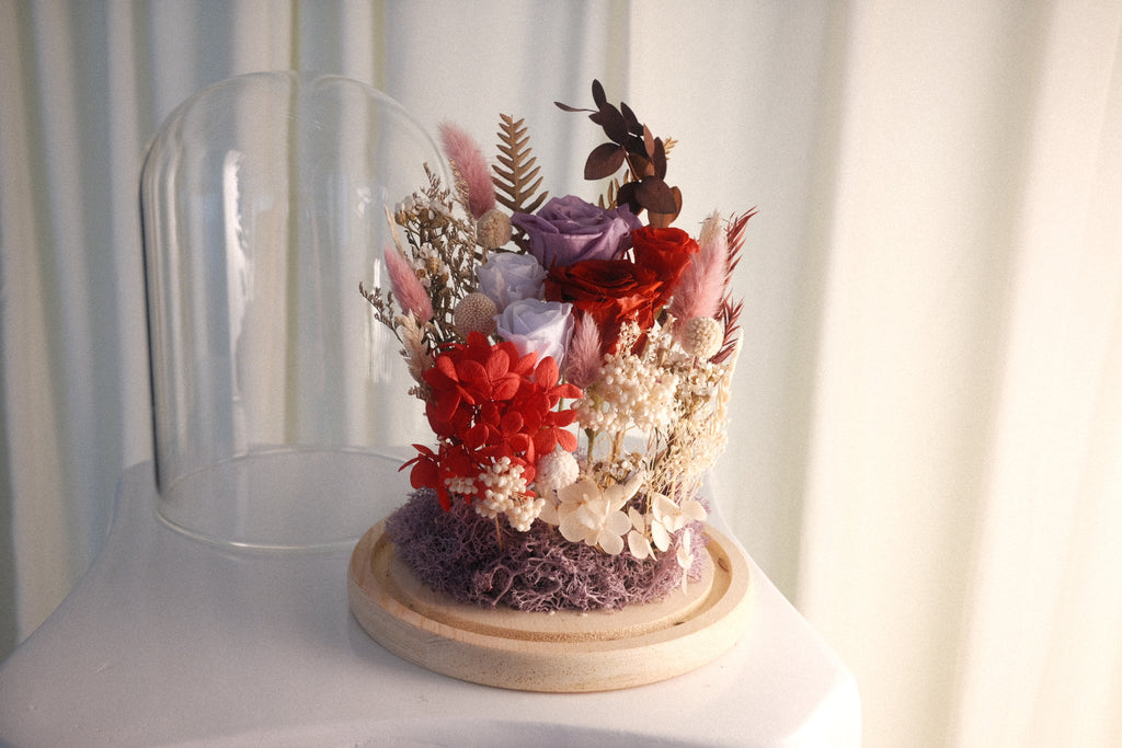 A preserved floral arrangement in a clear glass dome on a round wooden base, displayed on a white surface against a soft cream curtain background.