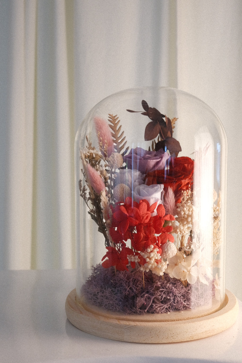 A preserved floral arrangement under a clear glass dome cloche on a round wooden base, set against a soft cream curtain background.
