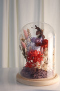 A preserved floral arrangement under a clear glass dome cloche on a round wooden base, set against a soft cream curtain background.