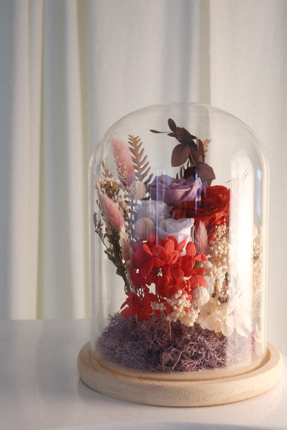 A preserved floral arrangement under a clear glass dome cloche on a round wooden base, set against a soft cream curtain background.
